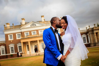 African wedding couple holding hands and cuddling outside the front of Boreham House. The groom, who is wearing a blue suit is kissing the bride on the cheek.
