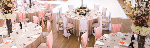 wedding reception set up with pink and white chair covers which surround several round tables and flower bouquets set up on table.