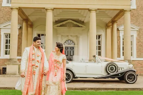 Asian wedding couple in the foreground smiling, standing outside the venue with a wedding car in the background